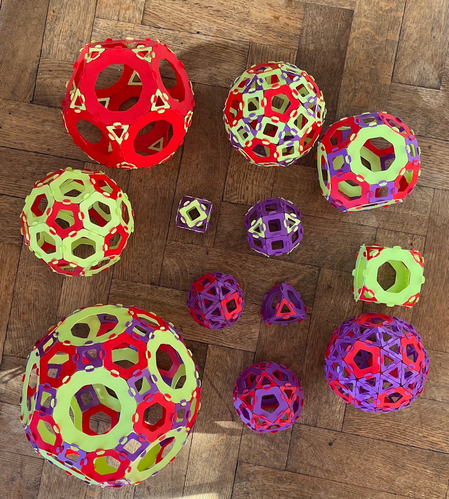 Une douzaine de solides géométriques en pièces rouges, violettes et vert clair, posés sur un parquet